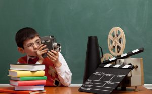 Little boy learning how to make film via video camera.He is filming via old fashioned camera in front of green blackboard.Stack of books, film slate,megaphone and film projector are on the desk.The blackboard is blank.He is wearing an orange waist and sitting on left side of frame.Indoor shot.